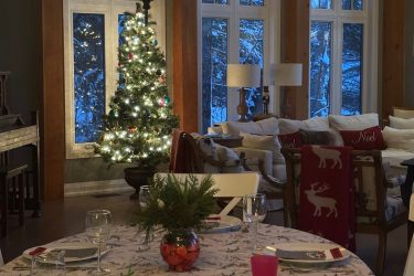 warm, rustic dining room and living space decorated for Christmas, featuring a lit Christmas tree by large windows, a oval dining table with festive place settings at muskoka soul luxury lakehouse rental